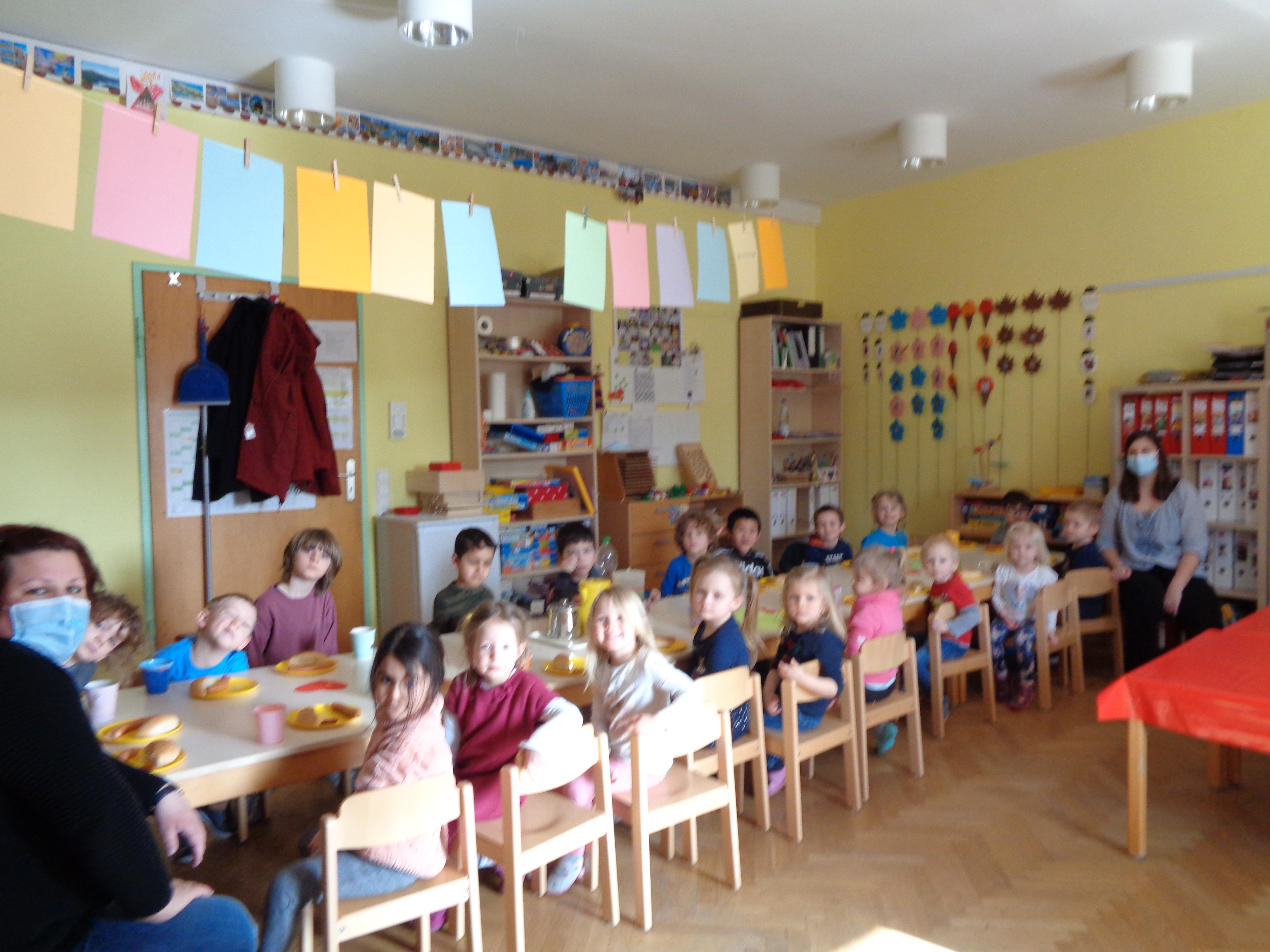  Hier sind Erziehende und Kinder zu sehen, wie sie an einem Tisch sitzend, einander willkommen heißen. 